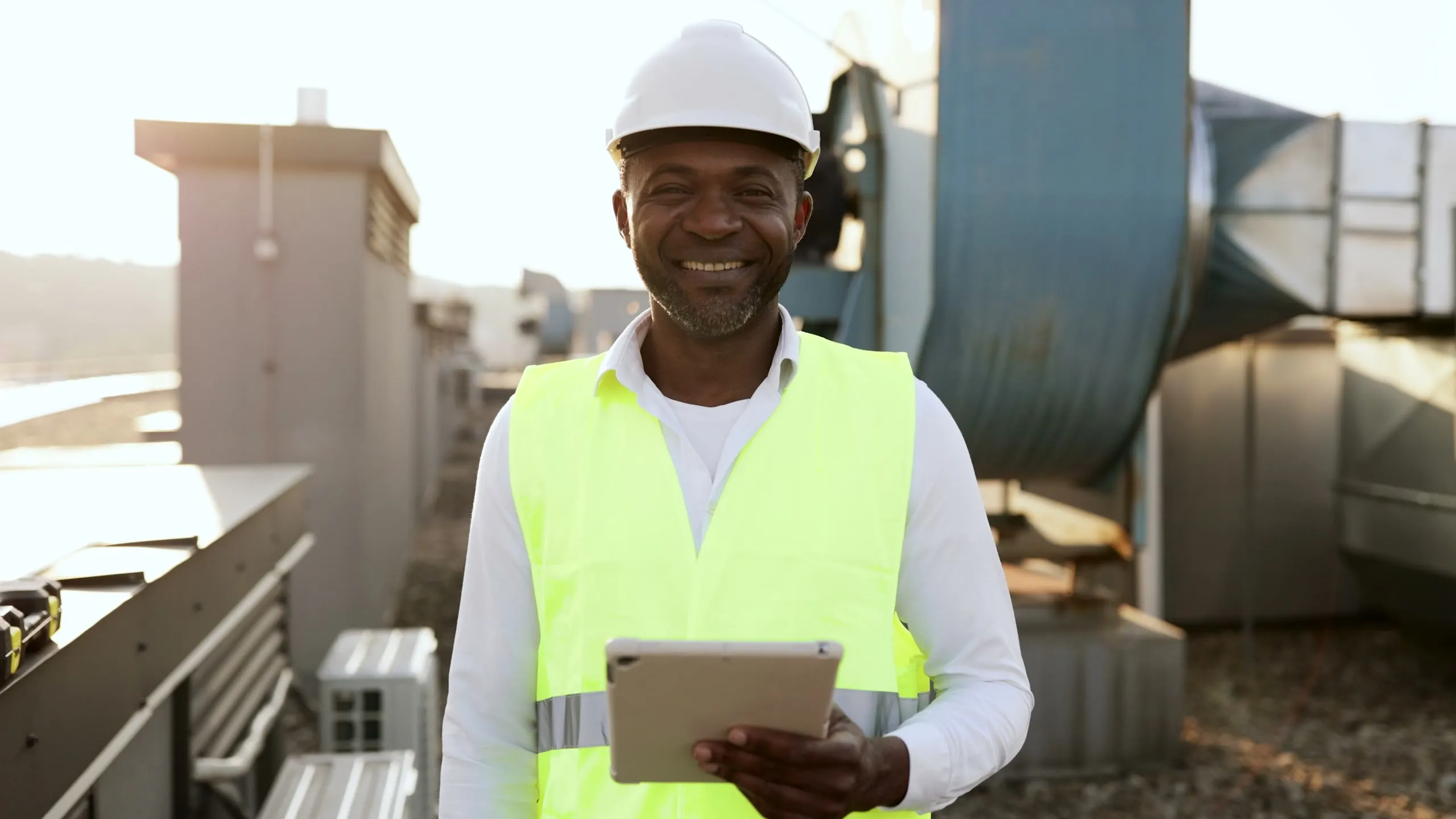 A technician in a hard hat walks across a building rooftop between HVAC units, using a tablet